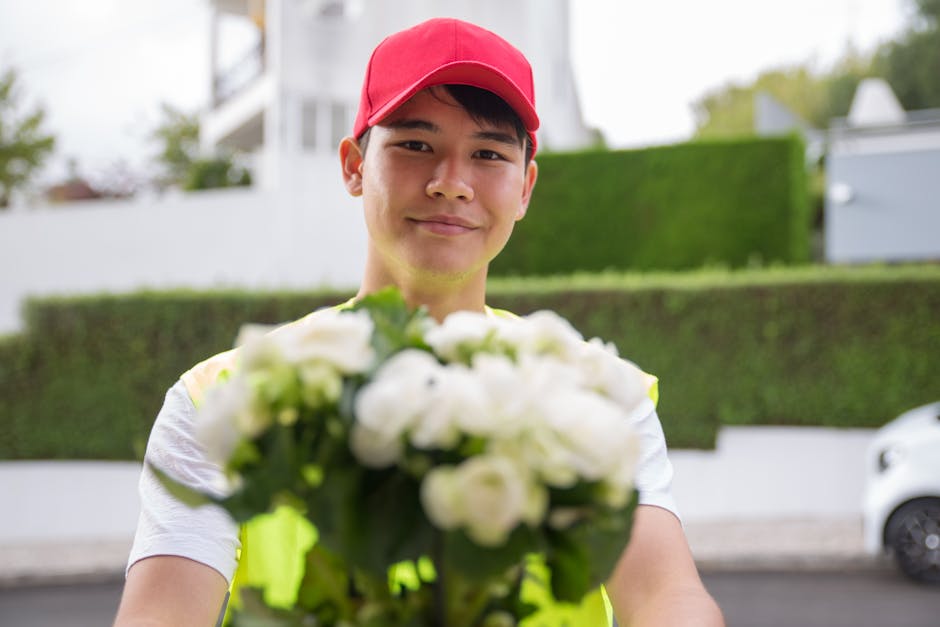 A delivery person wearing a red cap and high-visibility vest stands outdoors on a residential street with greenery and modern houses in the background, smiling and holding a bouquet of white roses and