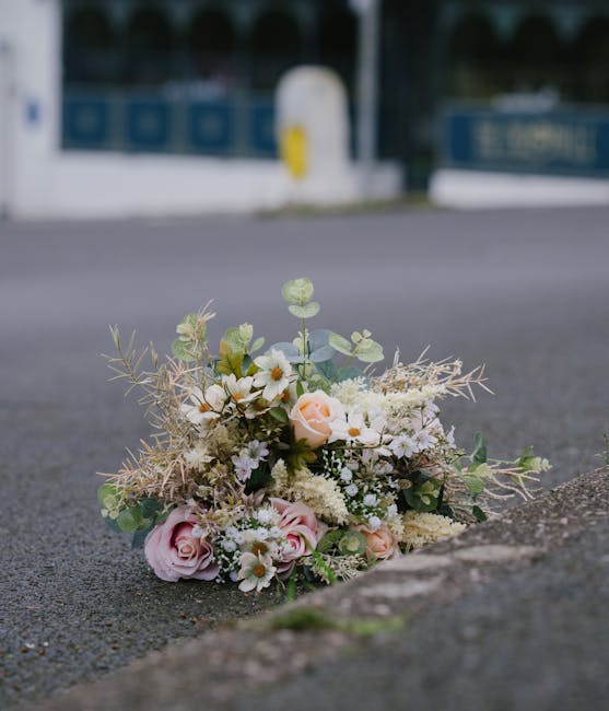A delicate floral bouquet placed on the pavement near the curb, featuring soft pink roses, white daisies, and small white filler flowers intertwined with muted green eucalyptus leaves and wispy beige