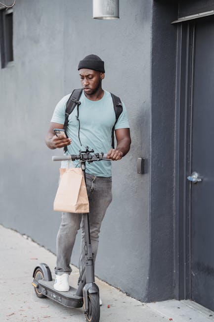 A young man with dark skin, wearing a grey beanie, light grey t-shirt, and grey jeans, stands on a grey sidewalk beside a grey wall and a closed grey door. He is holding and looking at a smartphone in