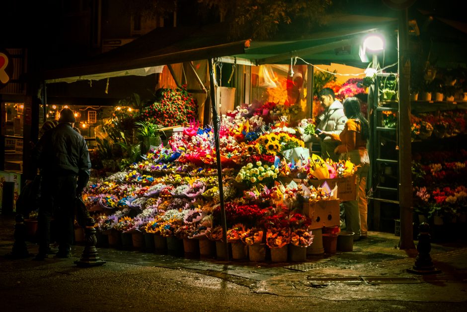 At a flower market stand illuminated by bright outdoor lighting, a vibrant display of fresh flowers is arranged in buckets and bouquets, featuring sunflowers, roses, lilies, and carnations in colors s