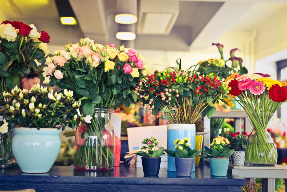 An arrangement of fresh flowers displayed on a dark wooden table inside a florist shop. The bouquet features pink and cream roses, white and pink carnations, and red and pink gerbera daisies, all arra