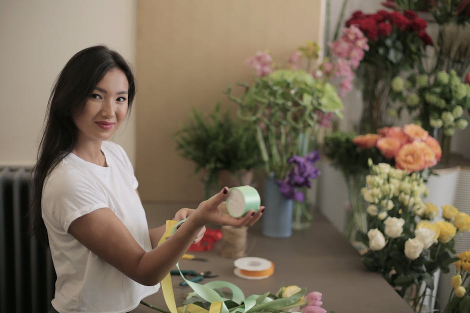 A young woman with long dark hair, wearing a white t-shirt, smiles gently while holding a roll of light green floral tape in her right hand and a piece of yellow ribbon in her left. She stands at a wo